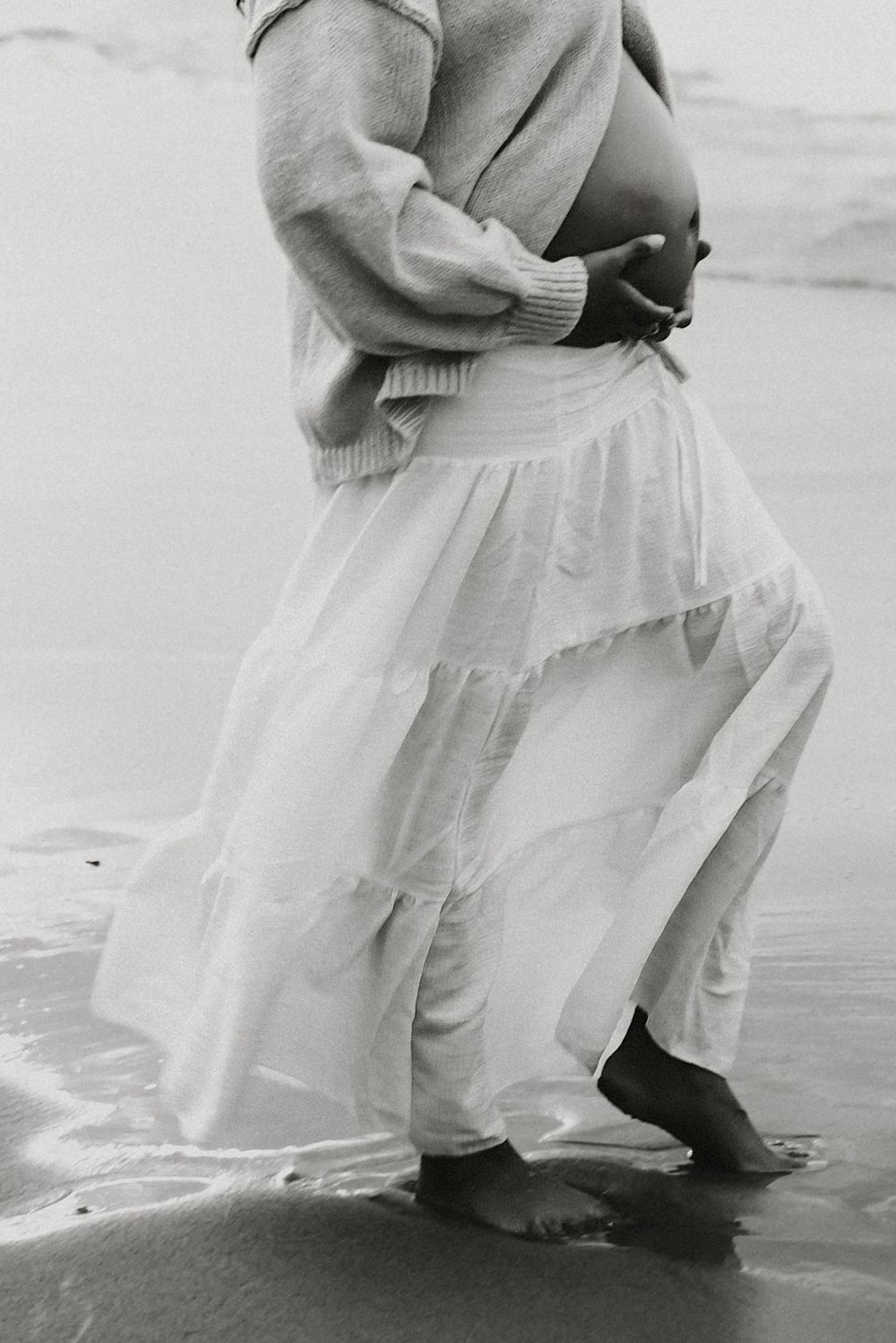 Black and white maternity photo of a pregnant woman in a knit sweater and flowing tiered skirt, barefoot on the wet sand by the ocean.