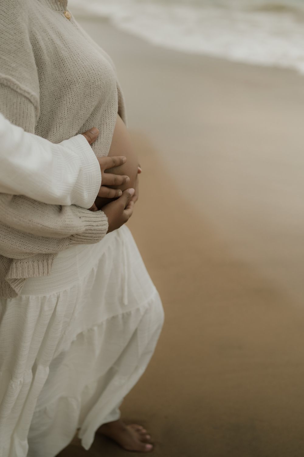 Pregnant woman in a cream knit sweater and white tiered skirt cradles her belly while standing barefoot on a sandy beach near the waves.