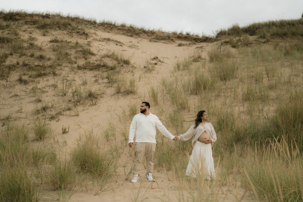 Couple holding hands in a sandy dune landscape with tall grass, he wears a white sweater and she a flowing cream dress.