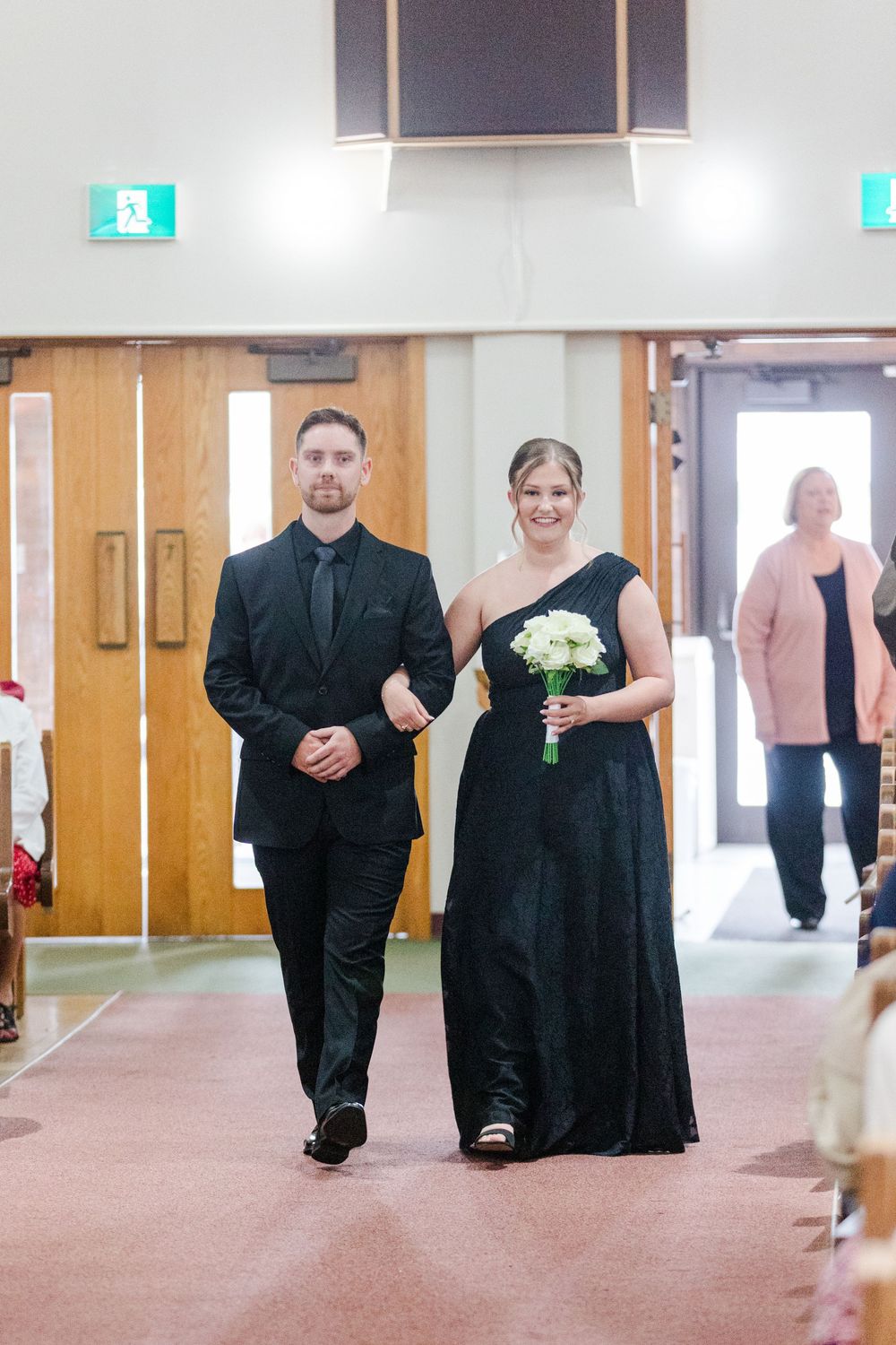 Groomsman in a black suit escorts a bridesmaid in a black gown holding a white bouquet down the church aisle during a wedding ceremony.