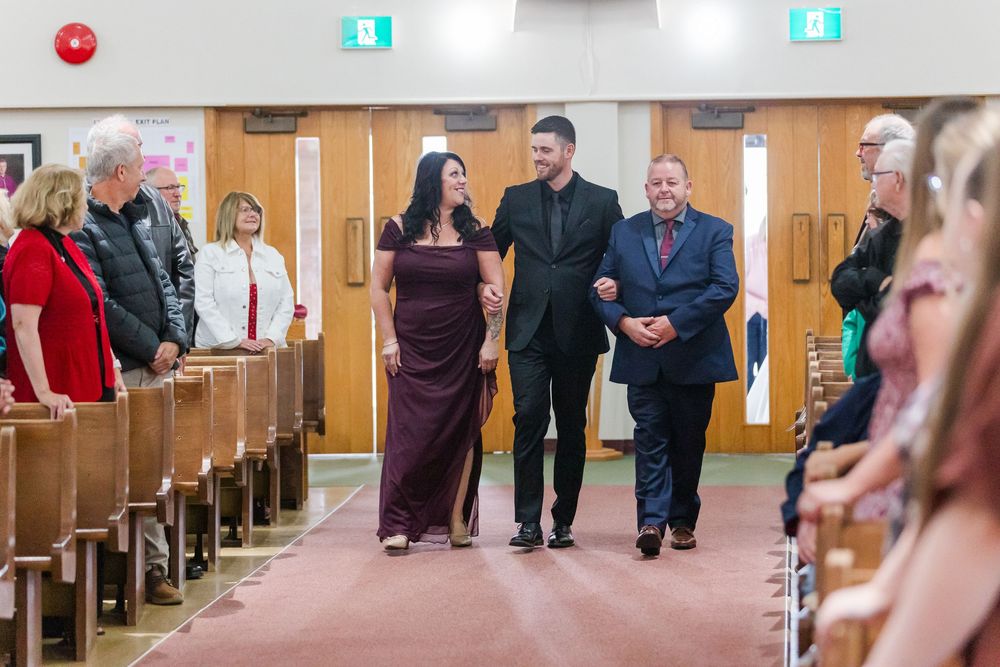 A woman in a burgundy dress walks arm in arm with two men in dark suits down a church aisle while guests watch from the pews.