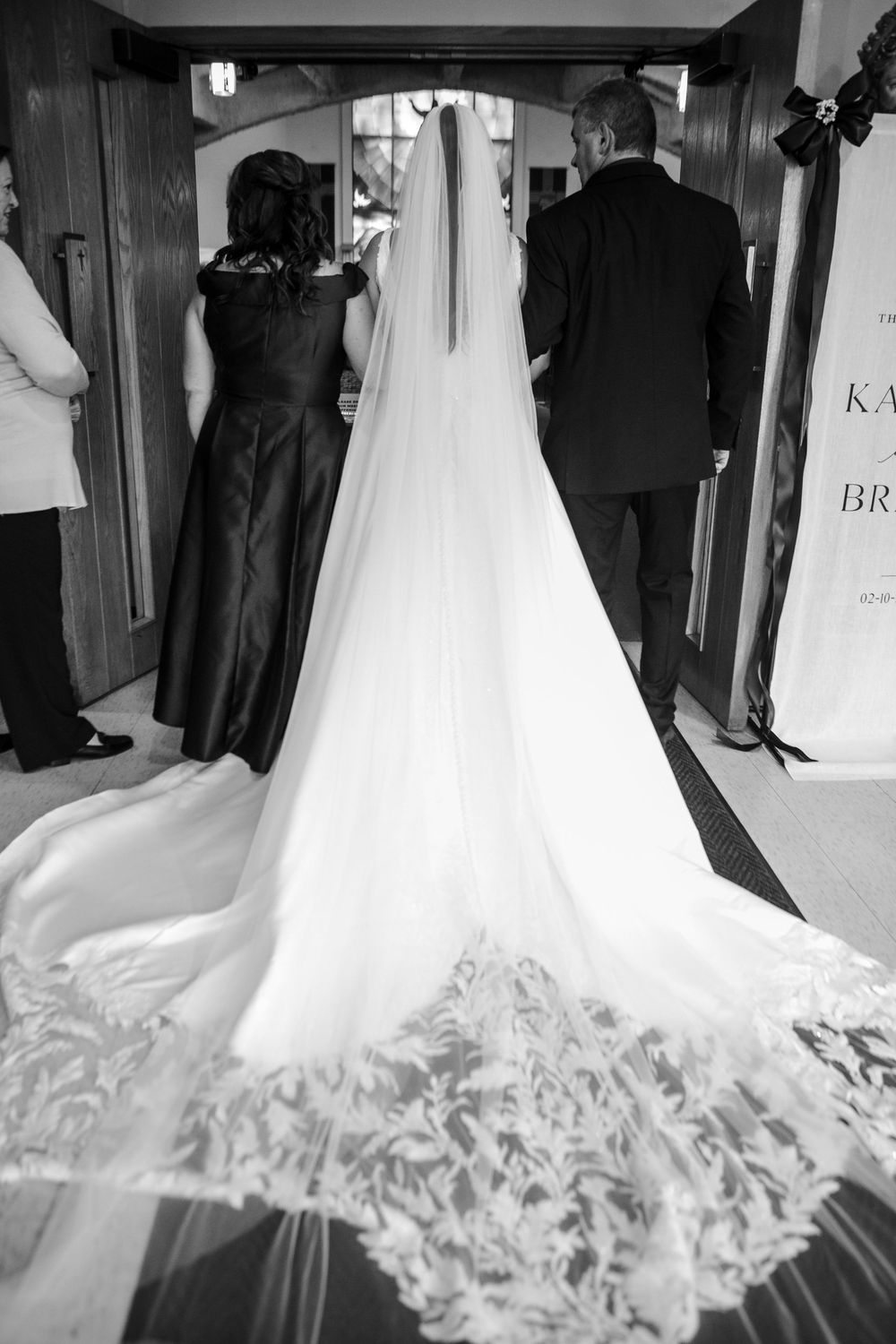 Black and white photo of a bride in a lace-trimmed gown with a long veil walking down the aisle with two guests.