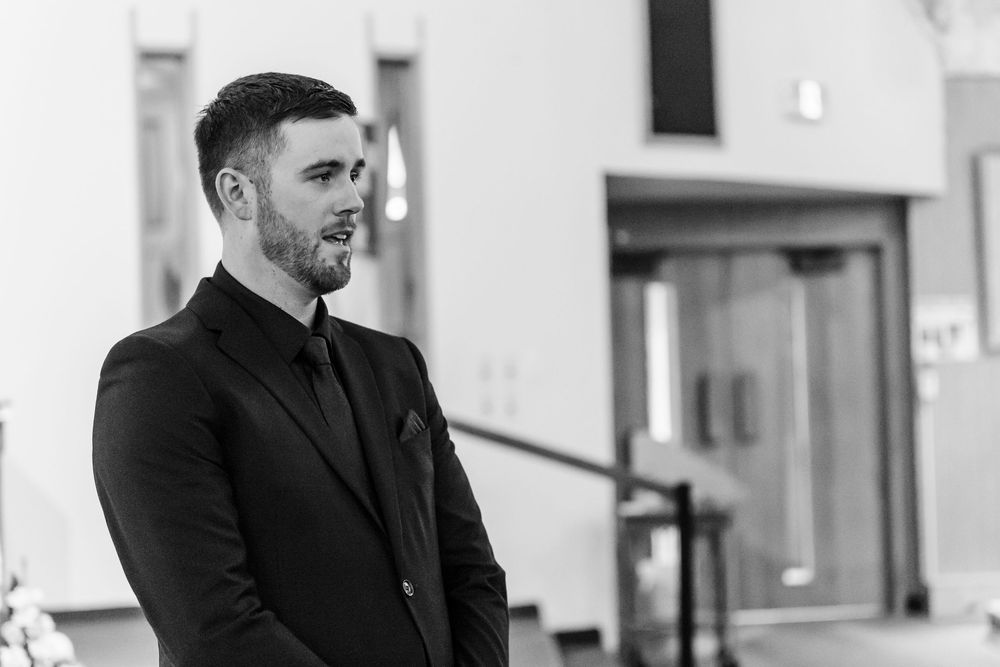 Black and white photo of a bearded man in a dark suit and tie standing in a church, gazing to the side with a calm, emotional expression.