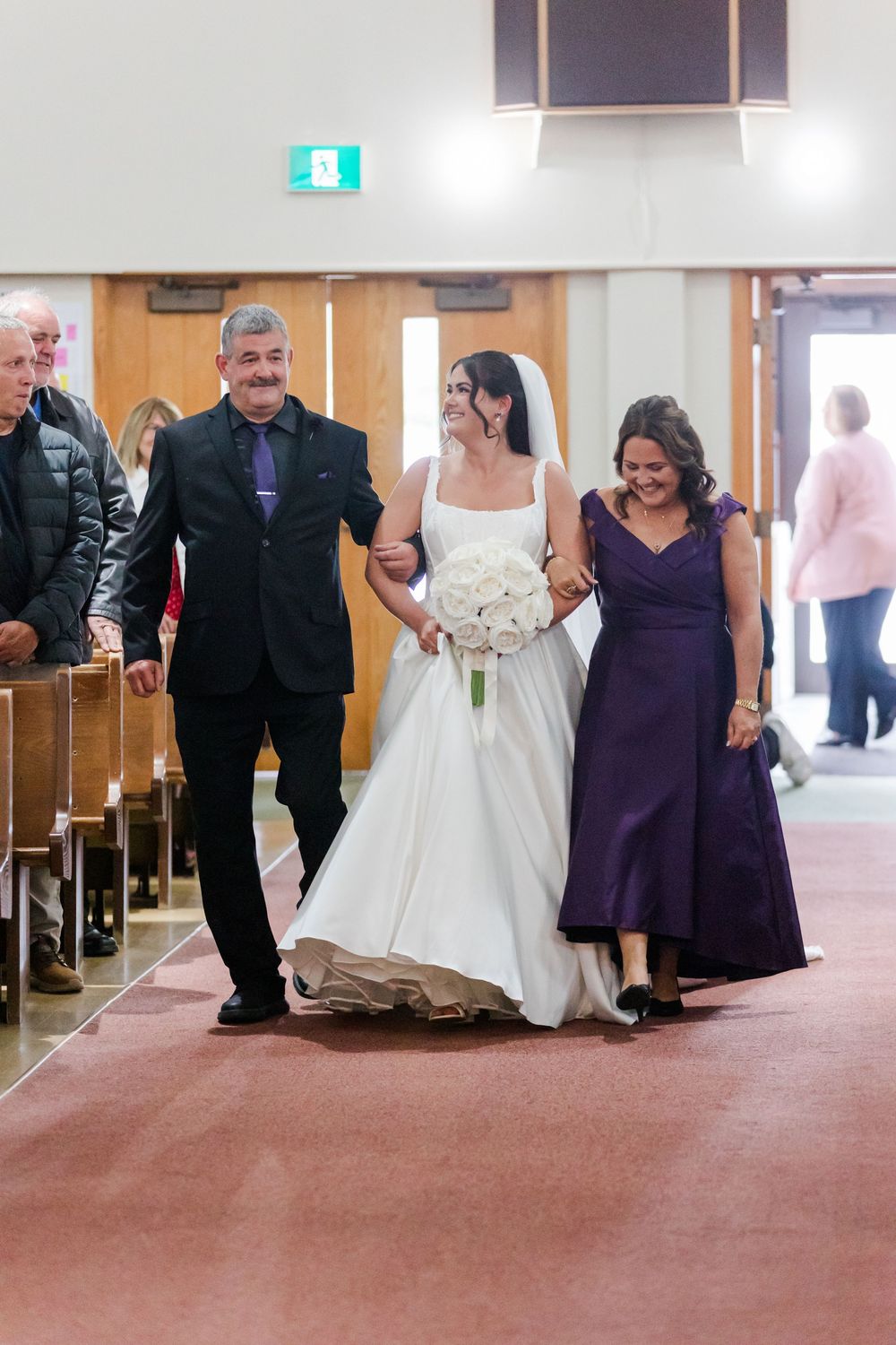 Bride in a white gown holding a bouquet walks down the church aisle arm in arm with a man in a dark suit and a woman in a purple dress.