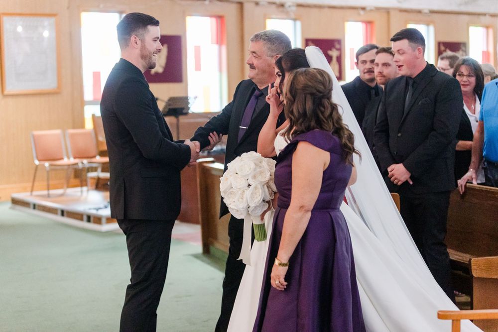 Father of the bride embraces the groom at the church altar as the bride in a white veil and bridesmaid in purple watch during the ceremony.