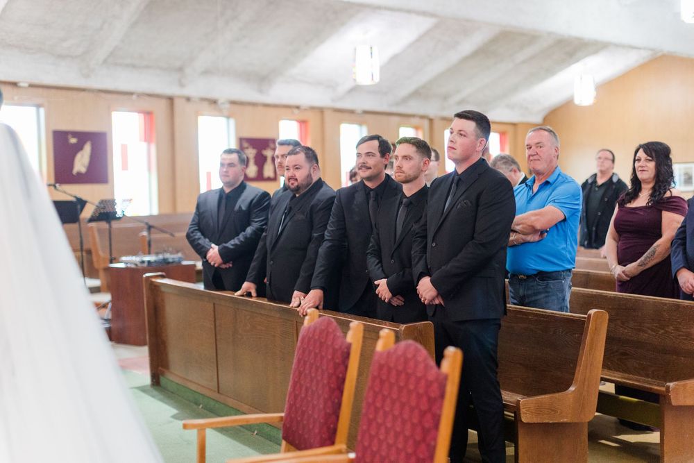 Group of men in dark suits standing beside wooden church pews during a ceremony, with other guests seated and standing nearby.