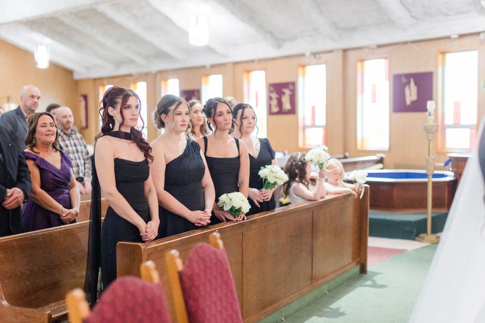 Bridesmaids in black dresses hold white bouquets while standing beside wooden church pews during a wedding ceremony as guests look on.