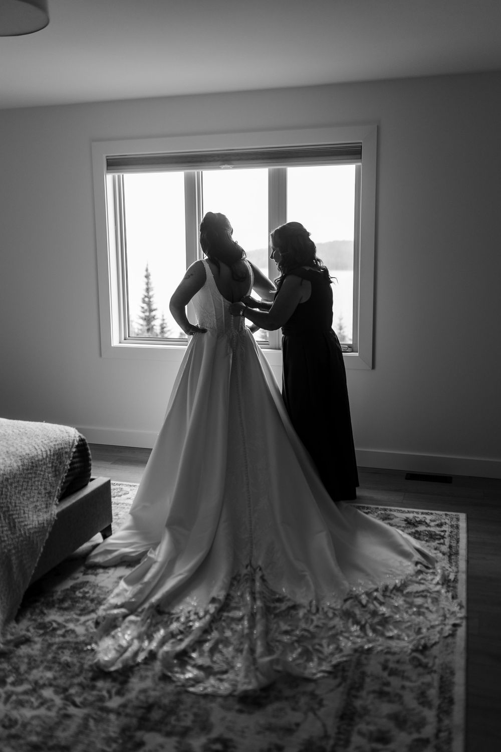 Black and white photo of a bridesmaid fastening the back of a bride's wedding gown with a long train beside a bright window.