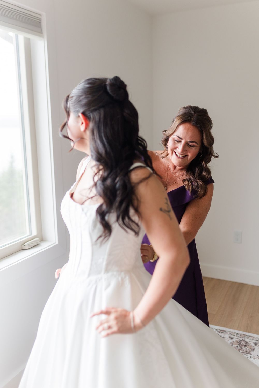 A smiling woman in a purple dress helps a bride in a white strapless wedding gown get ready beside a bright window.