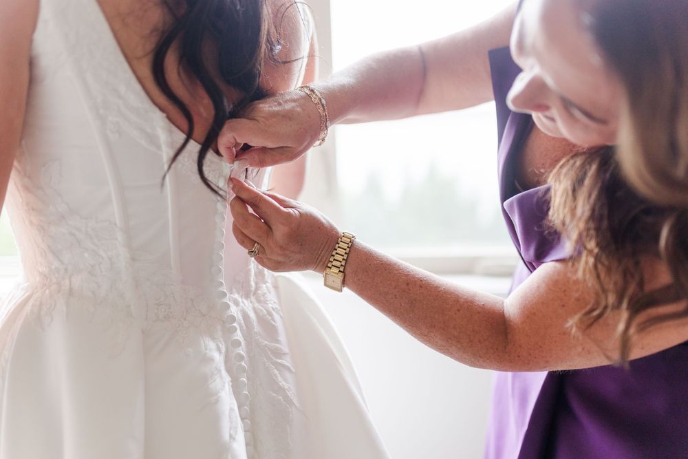 Woman in a purple dress helps a bride button the back of her white wedding gown in a bright room before the ceremony.