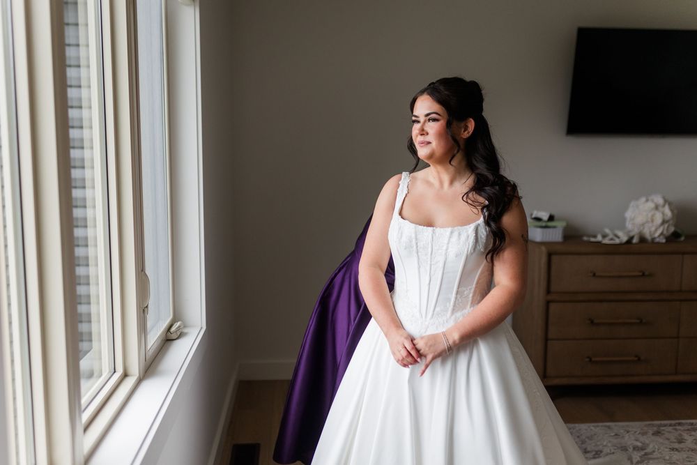 Bride in a white strapless wedding gown with dark hair pinned up gazes out a bright window beside a purple garment in a cozy bedroom.