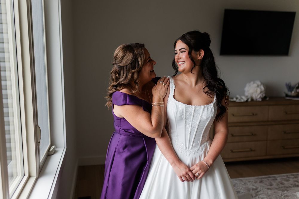 Bride in a white strapless gown smiles as her mother in a purple dress embraces her from behind in a bright bedroom before the wedding.