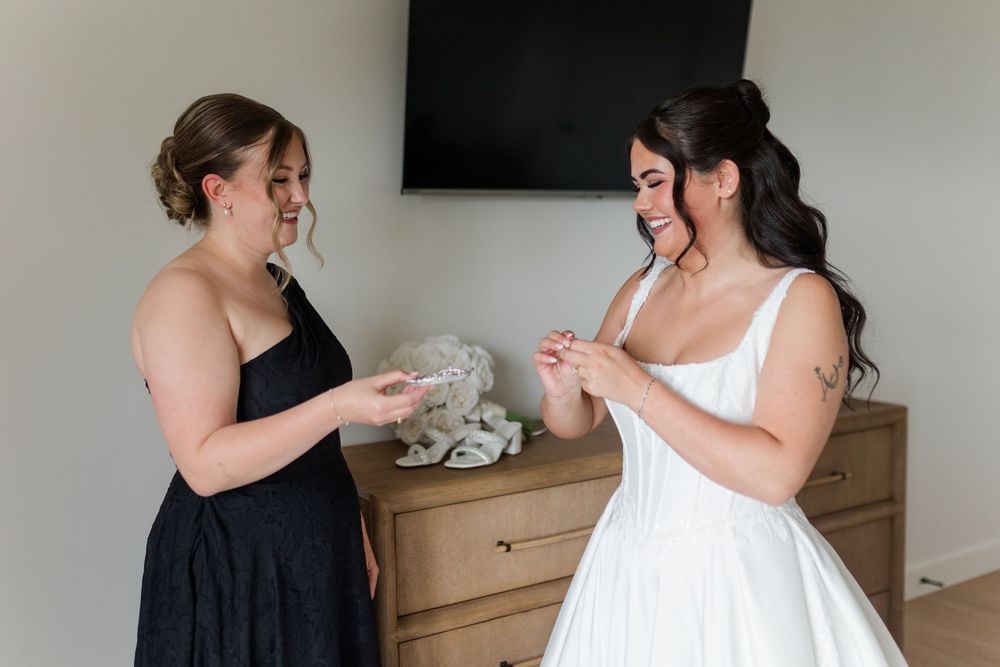 A bridesmaid in a black dress smiles while handing a small keepsake to a bride in a white gown in a bright bedroom.
