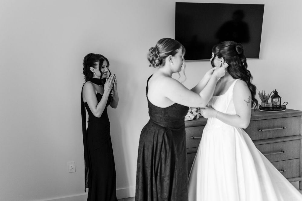 Black and white photo of a bride in a white gown being helped by a bridesmaid while another bridesmaid wipes away happy tears.