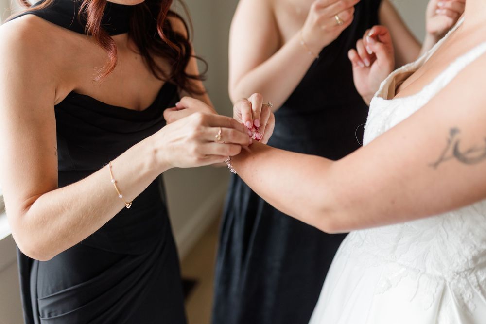 Bridesmaids in black dresses help a bride in a white gown fasten her bracelet during wedding day preparations.