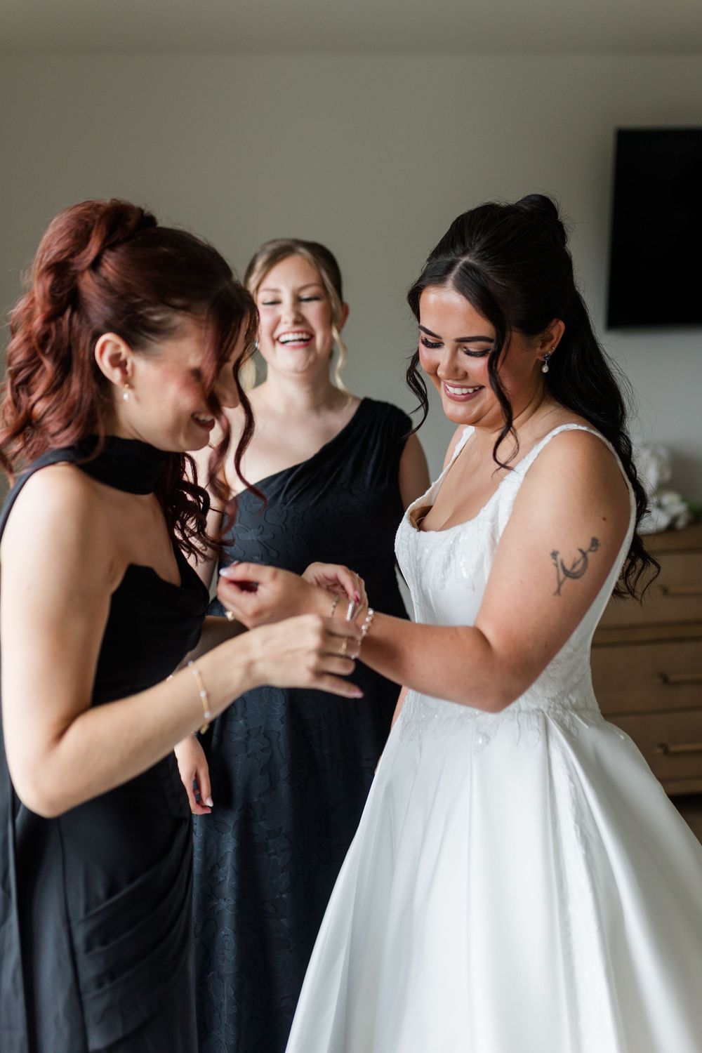 Smiling bride in a white strapless gown getting ready for her wedding with two bridesmaids in black dresses who help adjust her bracelet.