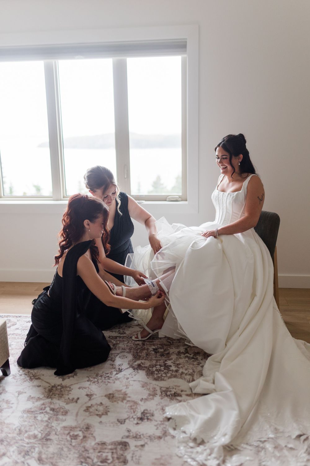 Two bridesmaids in black dresses help a smiling bride in a white gown put on her garter beside a bright window overlooking the water.