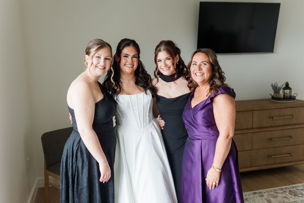 Bride in a white wedding gown poses with three smiling bridesmaids in black and purple dresses in a bright room.