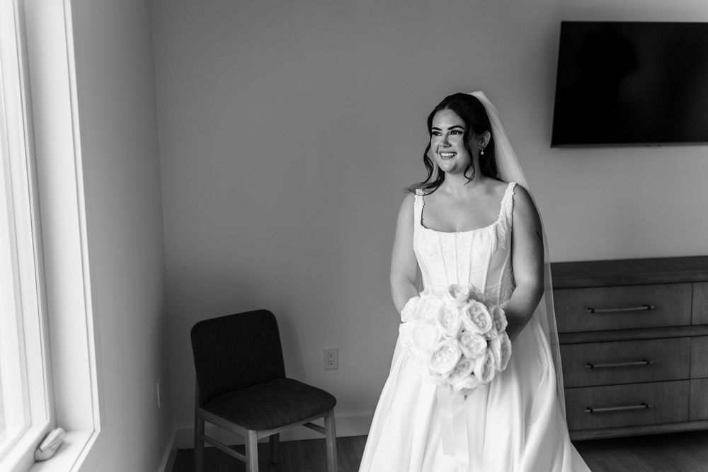 Black and white photo of a smiling bride in a white gown and veil holding a bouquet of roses beside a window in a bright room.