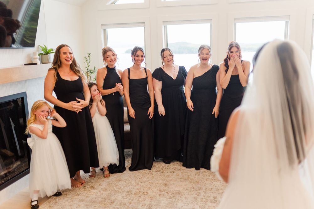 Bridesmaids in black dresses and a young flower girl react with joy as they see the bride in her veil during a first look.