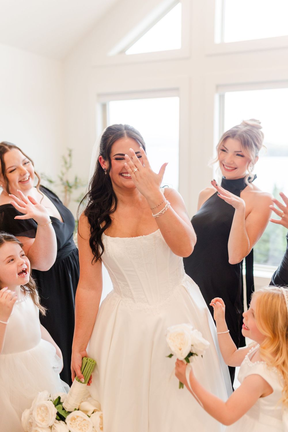 Smiling bride in a strapless white gown shows her ring while bridesmaids in black and flower girls with white bouquets celebrate.