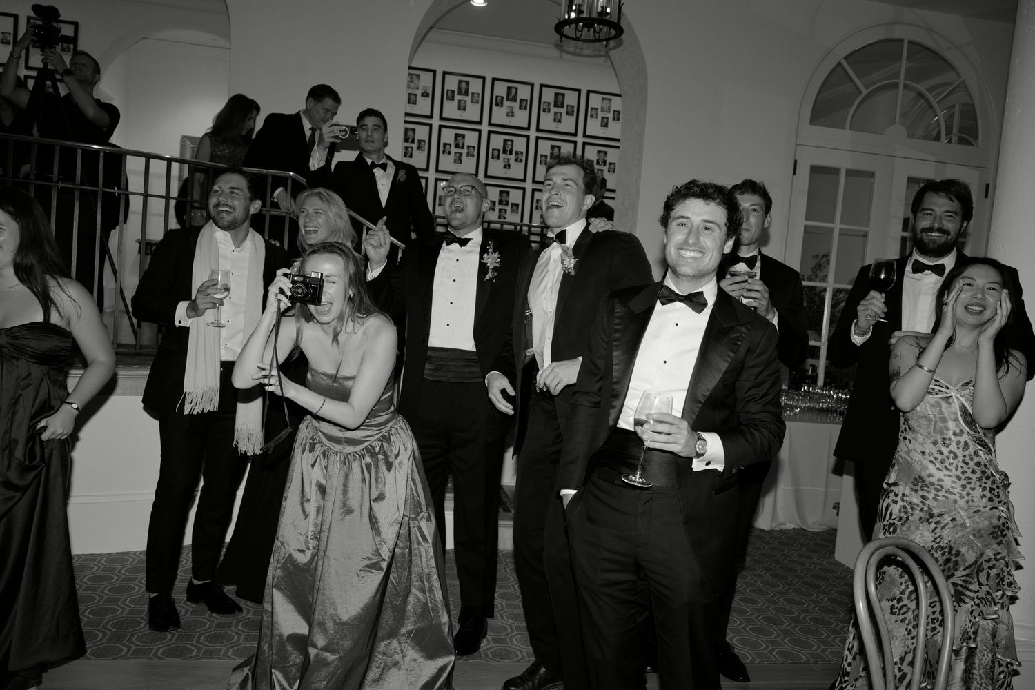Black and white photo of elegantly dressed guests in tuxedos and gowns socializing at a formal gala event.