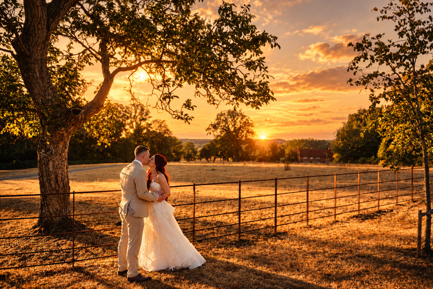Bride and groom share a romantic kiss at golden sunset by a fence and oak tree at an outdoor wedding venue.