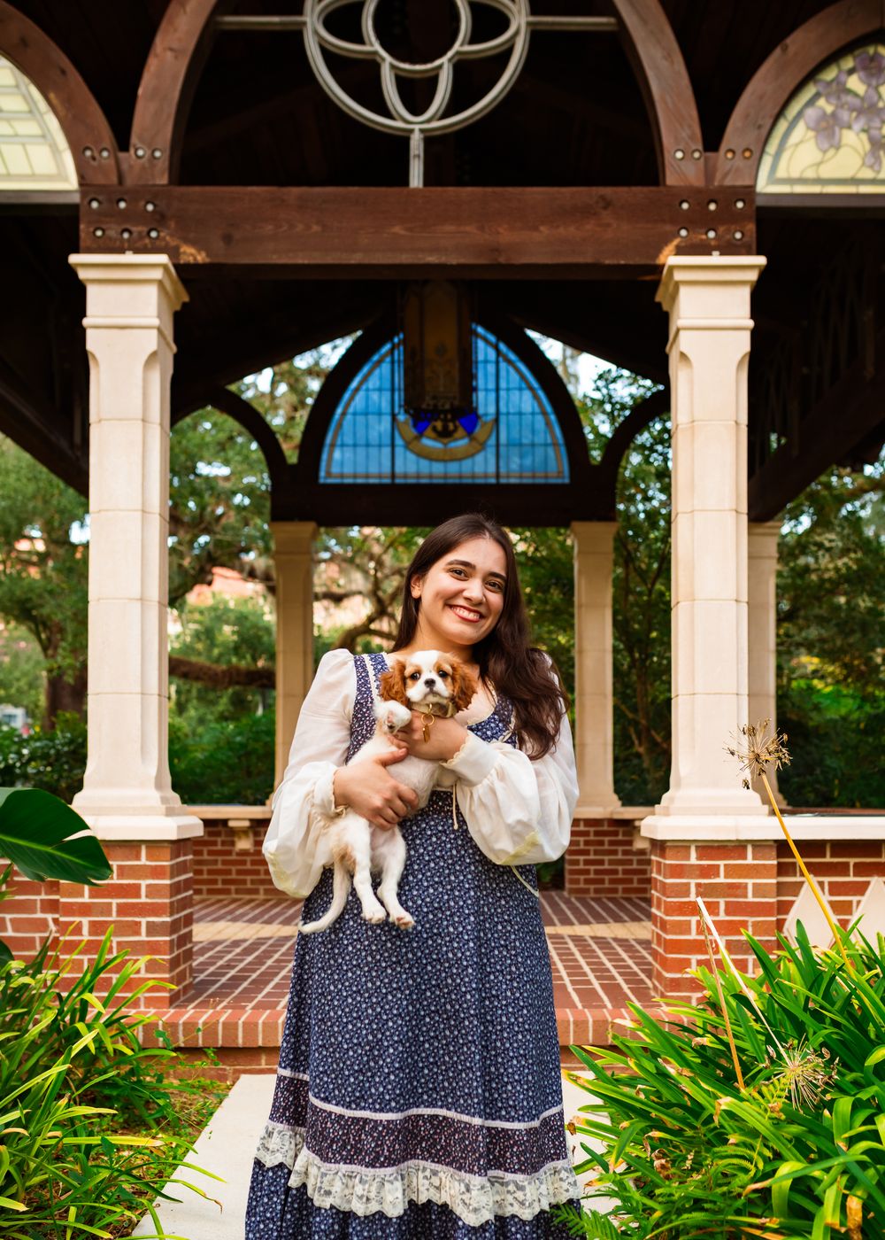 Young woman in floral dress holding a Cavalier King Charles Spaniel puppy in front of a brick gazebo.