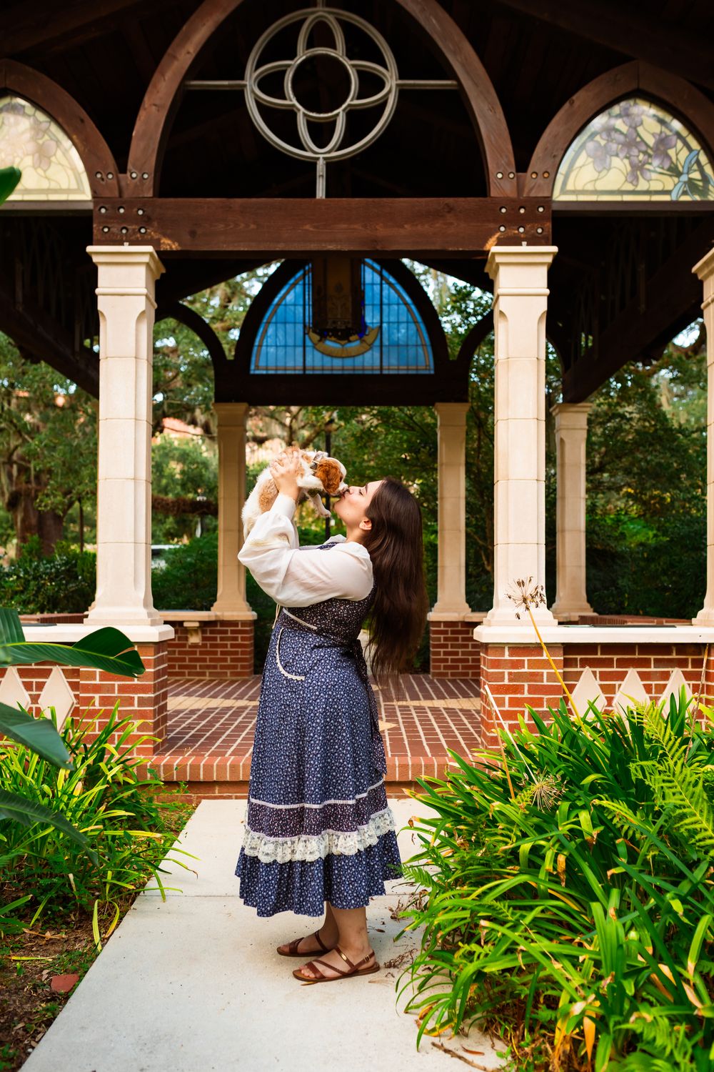 Young woman in blue floral dress kisses small puppy while standing in elegant brick gazebo surrounded by lush greenery.