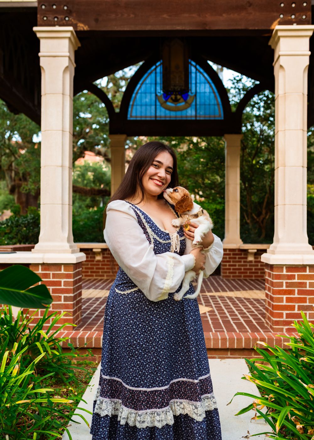 Young woman in blue floral dress holds a small dog, smiling in front of a brick gazebo with stained glass.