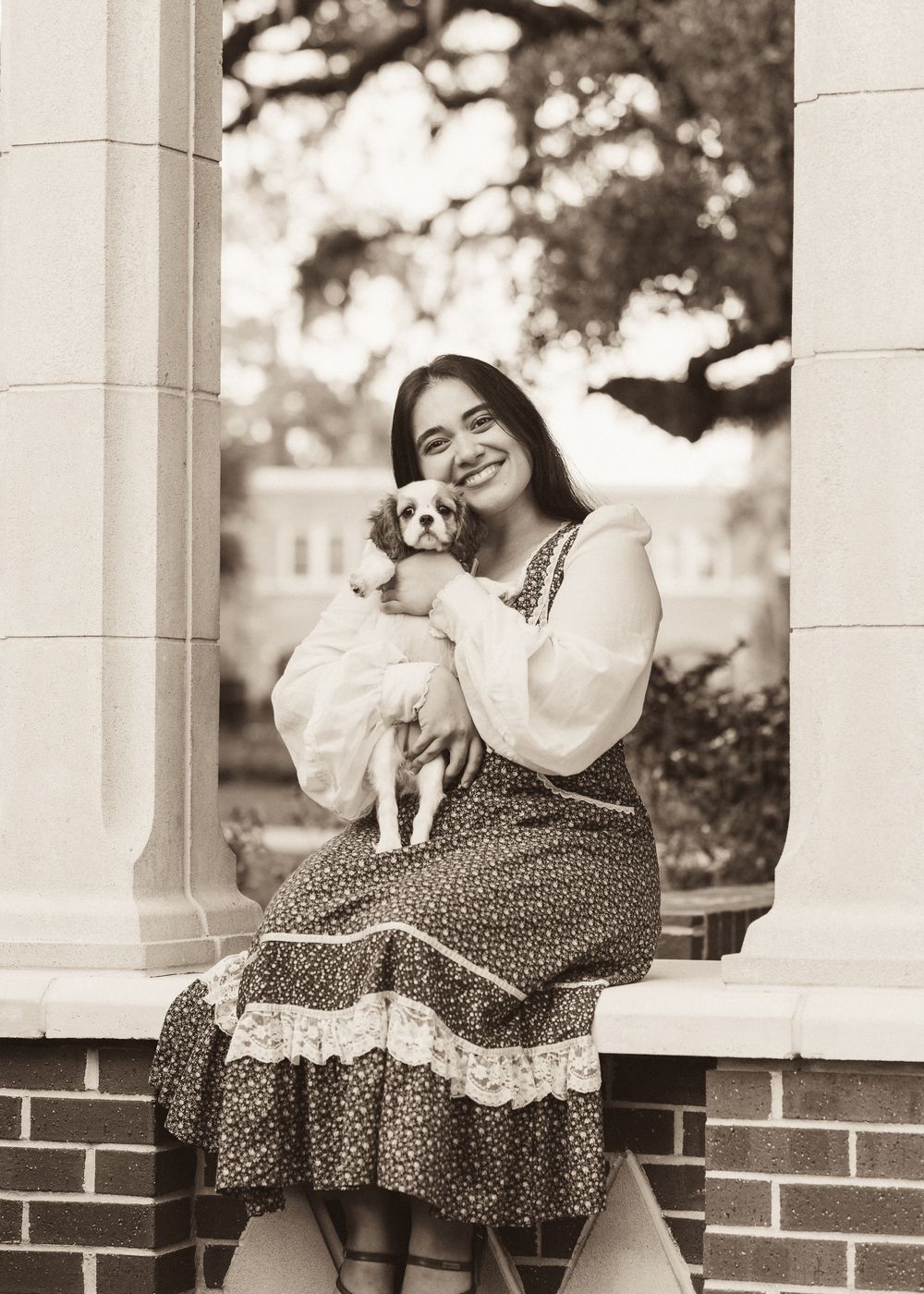 Smiling woman in floral dress holding small white dog, seated on brick ledge, sepia-toned portrait photo.