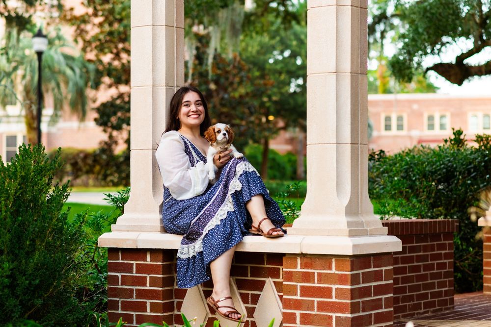 Young woman sitting on brick columns with a small Cavalier King Charles Spaniel dog, posing outdoors near a classic building.