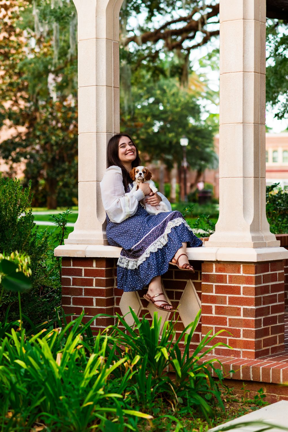 Young woman in blue floral dress holding small puppy while sitting on brick ledge in garden gazebo.