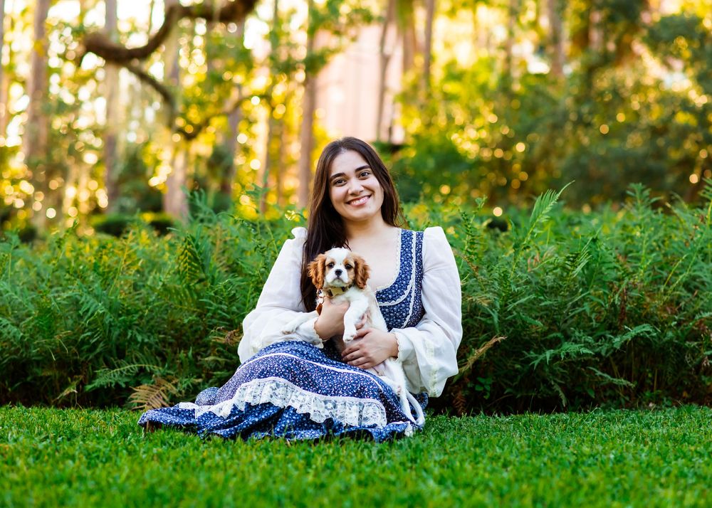 Young woman in blue floral dress sits on green grass holding a Cavalier King Charles Spaniel puppy in a lush garden.