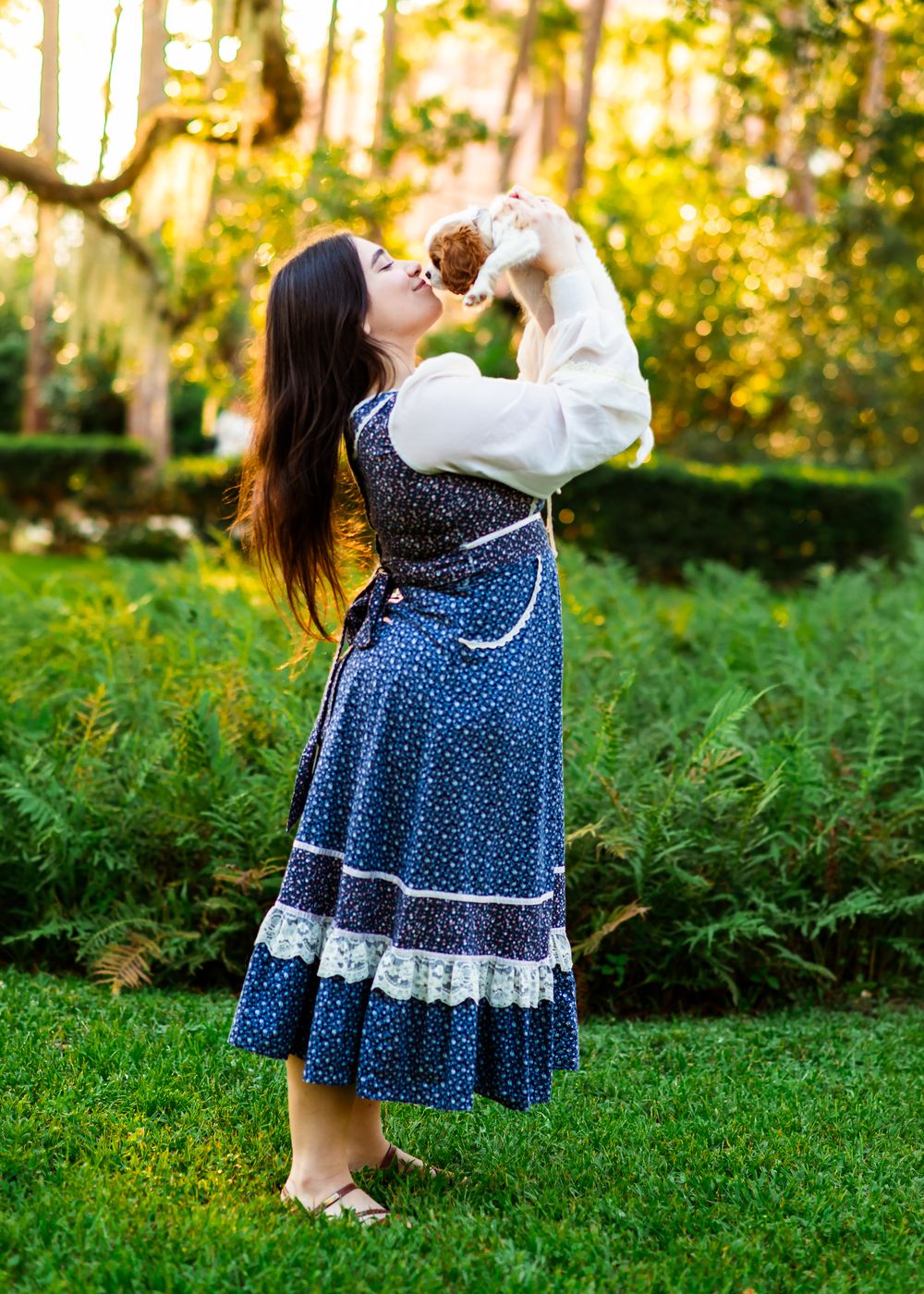 Woman in blue polka dot dress with lace trim kisses small puppy outdoors in lush green garden.
