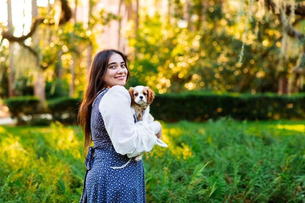 Young woman in polka dot dress holds small dog in lush green park, smiling over shoulder in golden sunlight.