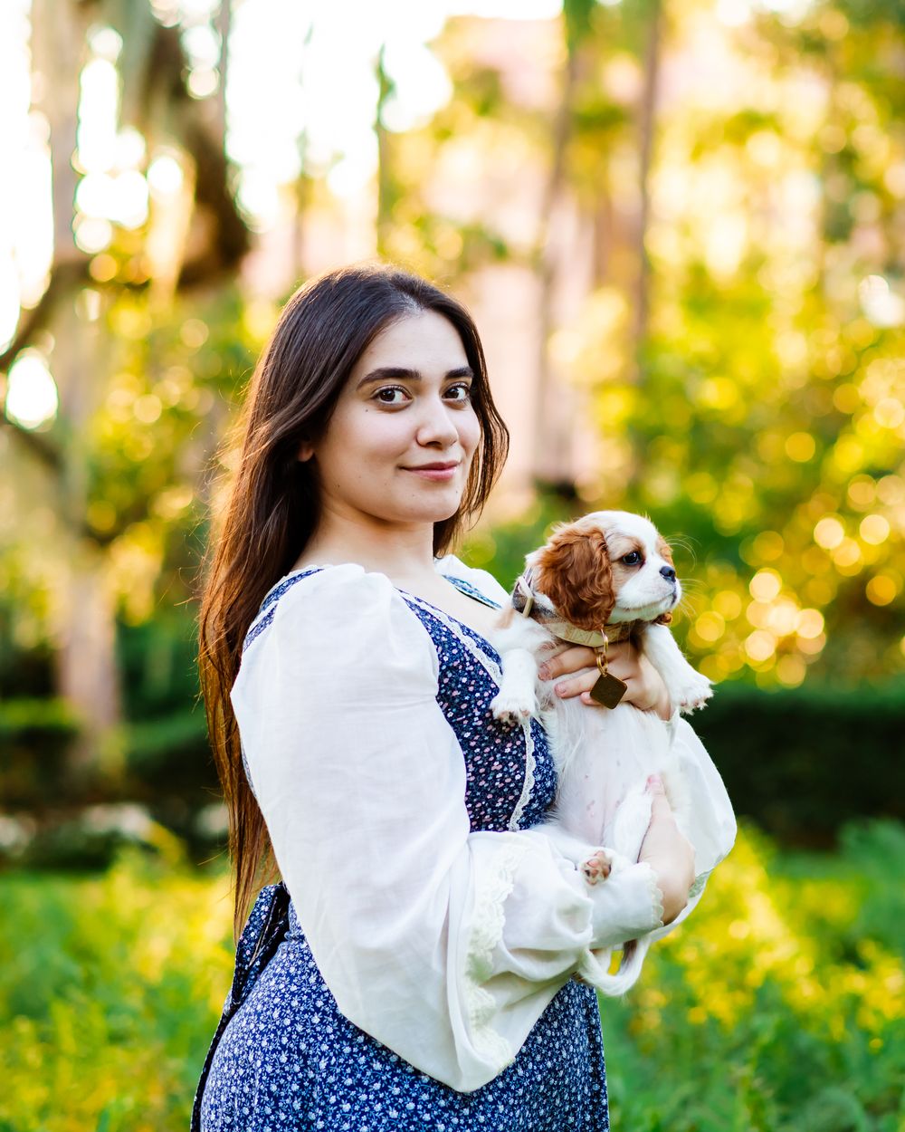 Smiling young woman with long dark hair holds adorable Cavalier King Charles Spaniel puppy outdoors in golden light.