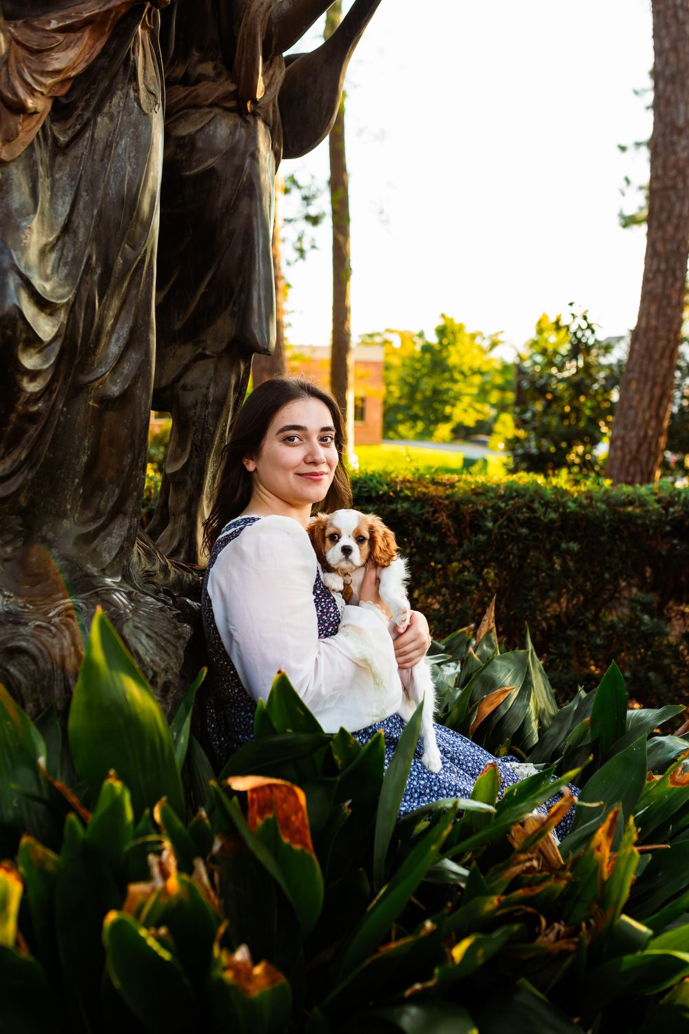 Young woman in white blouse holds a Cavalier King Charles Spaniel puppy beside a bronze statue in a lush garden.
