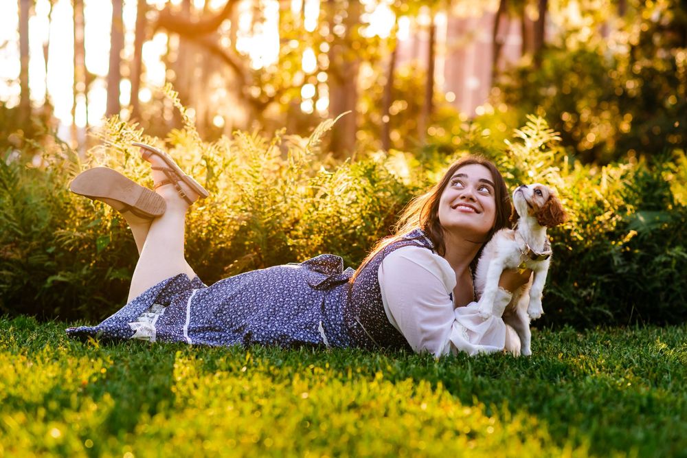 Young woman lying on green grass smiling at her small dog in a sunny golden-hour garden setting.