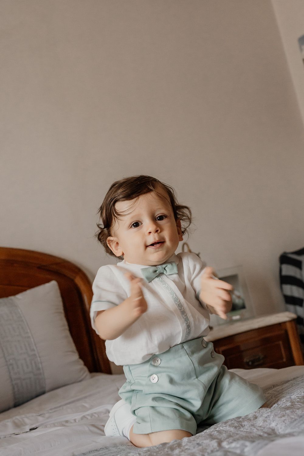 Adorable toddler in white shirt and mint green shorts sitting on a bed, clapping hands and smiling.