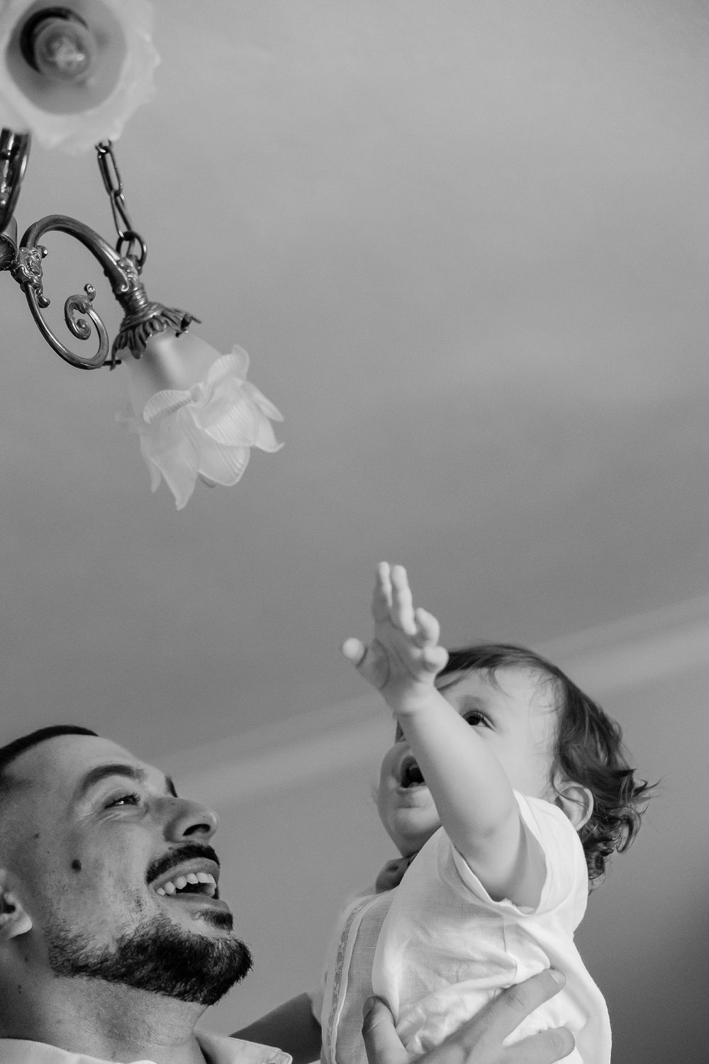 Black and white photo of a laughing father lifting his baby toward a chandelier light fixture indoors.