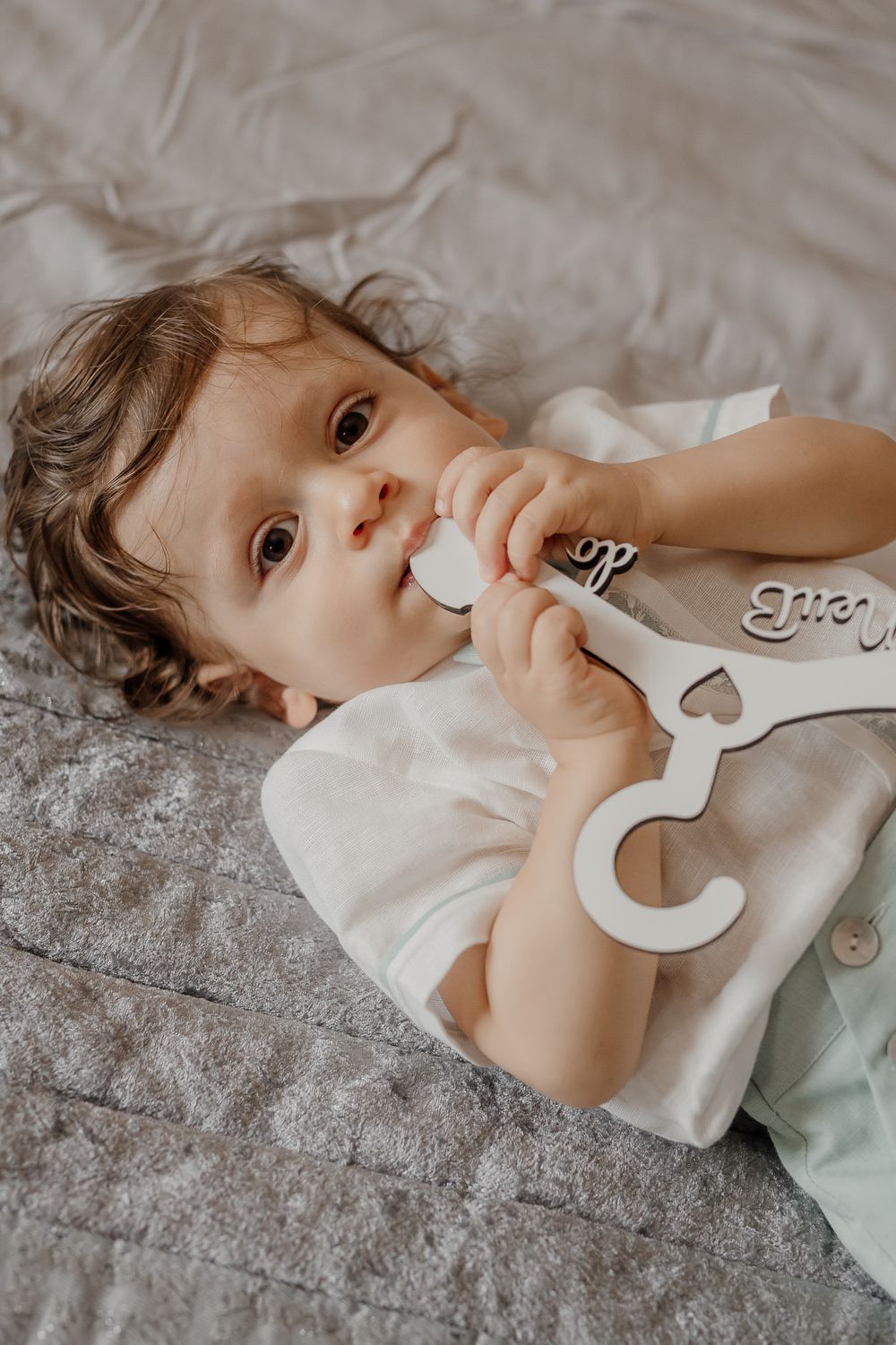 A cute baby lying on a gray bed, holding a white wooden name hanger with cursive lettering.