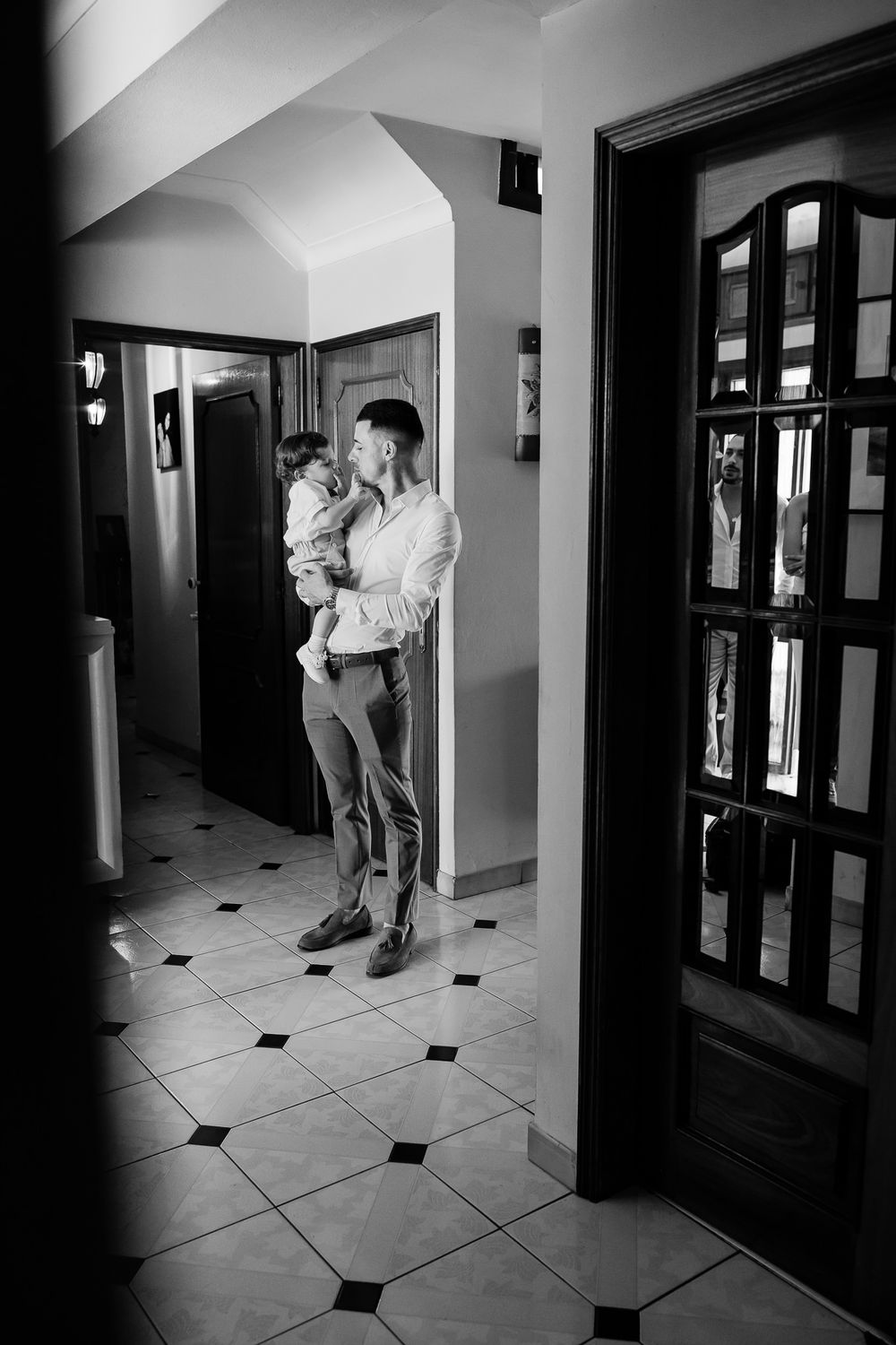Black and white photo of a man holding a young child in an elegant hallway with tiled floors and wooden doors.