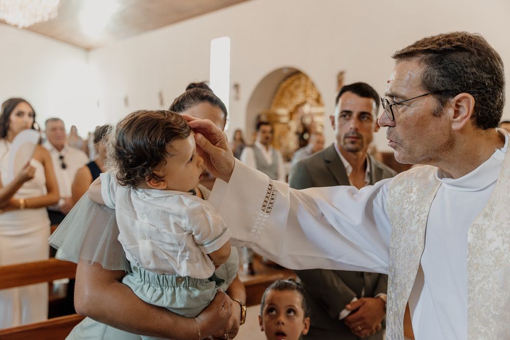 A priest anoints a baby during a baptism ceremony inside a church, with family members watching in the background.