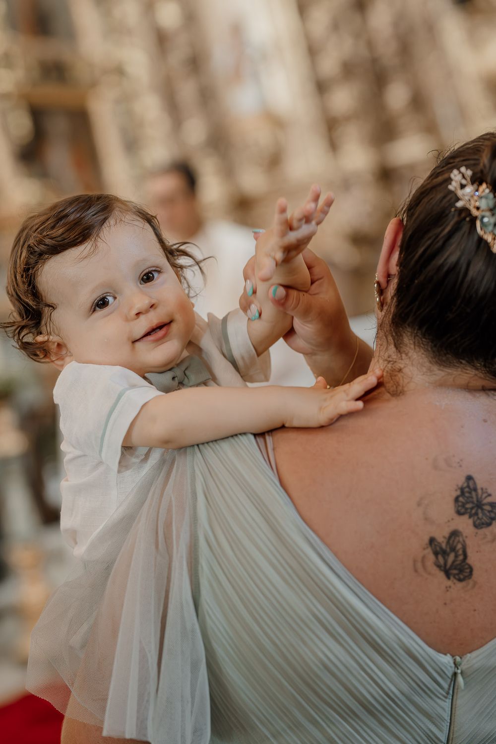 A baby in a white outfit reaches toward a woman with butterfly tattoos at an elegant event.