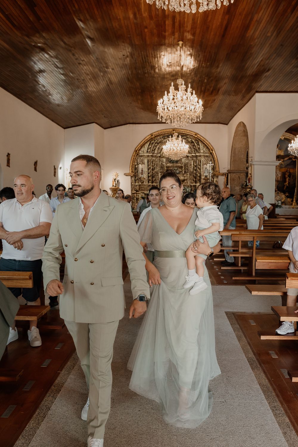 Couple walks down church aisle, man in beige suit, woman in sage green gown holding baby, crystal chandelier above.