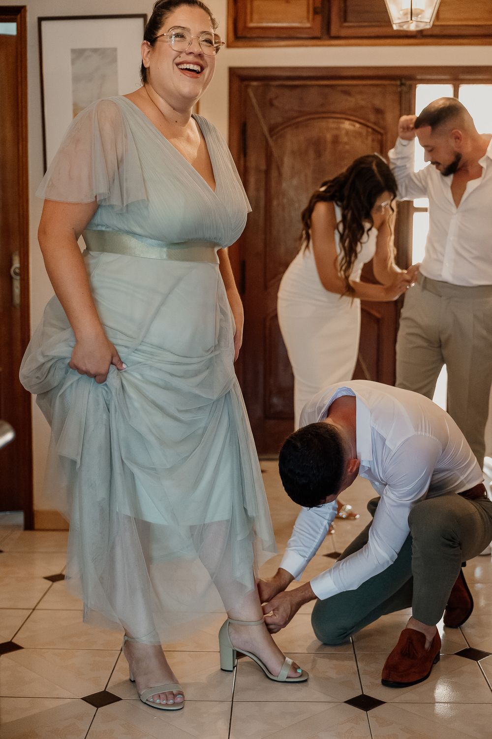 A bridesmaid in a sage green dress laughs as someone helps fasten her heels, with bride and groomsman in background.