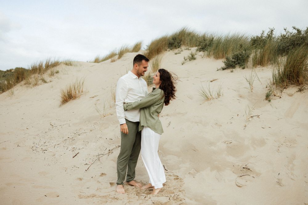 Couple embracing on sandy beach dunes, woman in white dress and man in sage green shirt, romantic outdoor photoshoot.