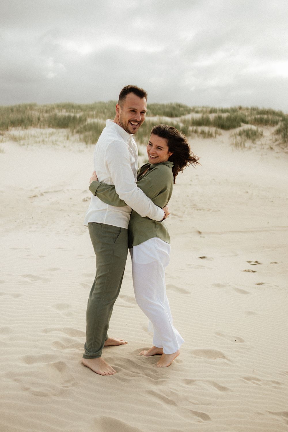 A happy couple embraces barefoot on a sandy beach, wearing casual green and white outfits, with grass-covered dunes behind them.