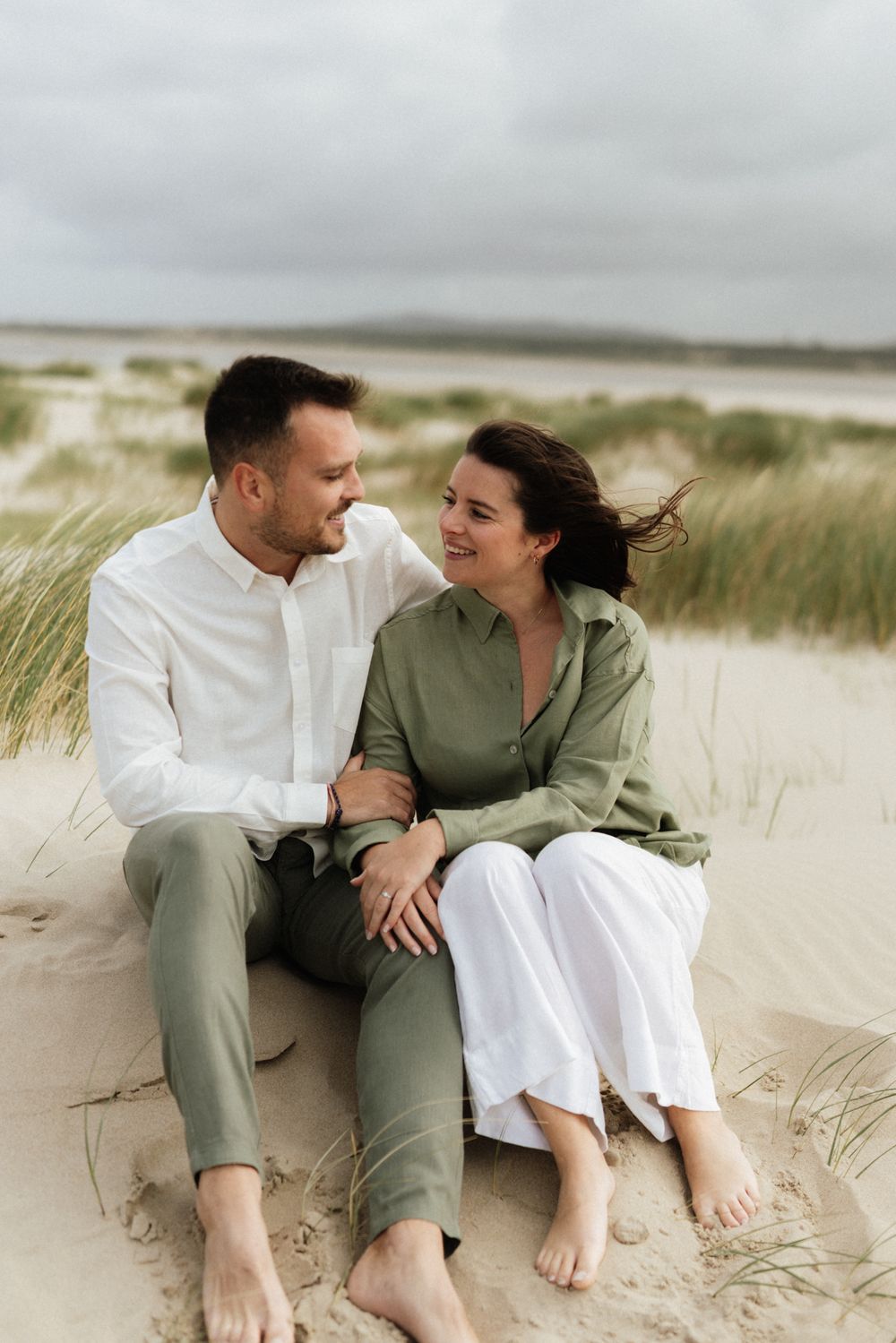 A couple sits barefoot on sandy beach dunes, smiling at each other, dressed in green and white linen outfits.